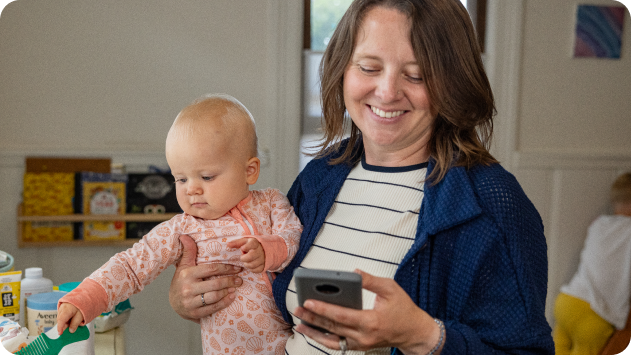 A parent holding a baby with one hand and looking at their cell phone in their other hand.