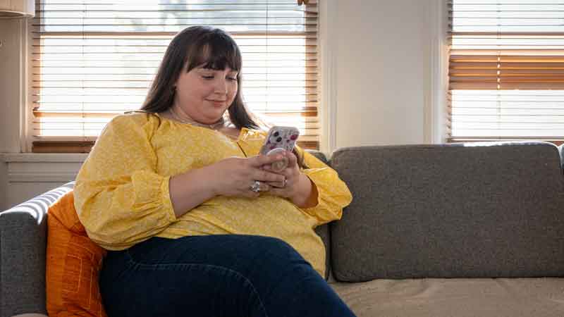 A woman sits on a couch while looking at a smart device.